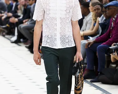 Male model in a white lace shirt and dark trousers walks a fashion runway, holding a patterned bag.