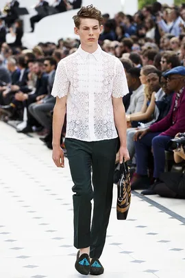 Male model in a white lace shirt and dark trousers walks a fashion runway, holding a patterned bag.