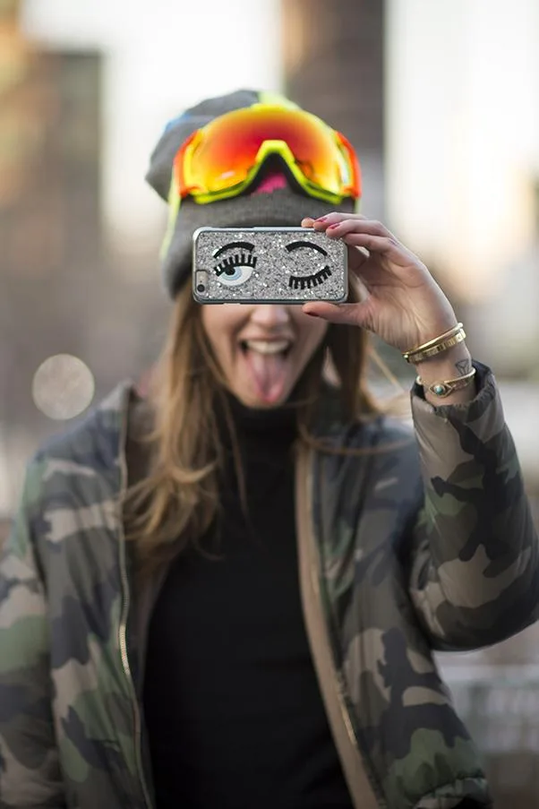 A woman in a camo jacket and goggles sticks her tongue out and holds a phone with a winking face case in front of her face.