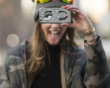 A woman in a camo jacket and goggles sticks her tongue out and holds a phone with a winking face case in front of her face.