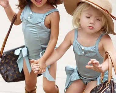 Two young girls in matching blue outfits, large tan hats, and sandals, each holding a designer bag while standing on a pavement.