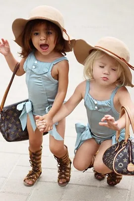 Two young girls in matching blue outfits, large tan hats, and sandals, each holding a designer bag while standing on a pavement.