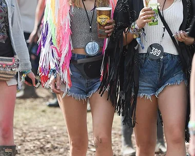 Two women in festival attire, wearing colorful fringed jackets and sunglasses, holding drinks at an outdoor event.