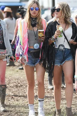 Two women in festival attire, wearing colorful fringed jackets and sunglasses, holding drinks at an outdoor event.