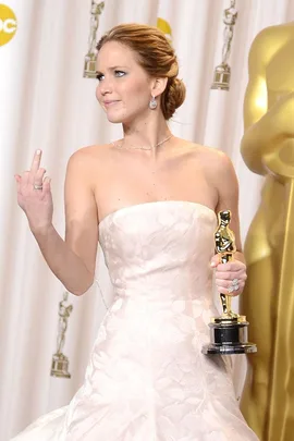 Woman in white strapless dress holding an Oscar, making a defiant gesture with her middle finger.