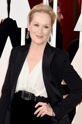 Meryl Streep poses in a black blazer and white blouse at an event, smiling warmly with her hand on her hip.