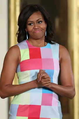 Michelle Obama speaking to high school students while wearing a colorful checkered dress and blue earrings.