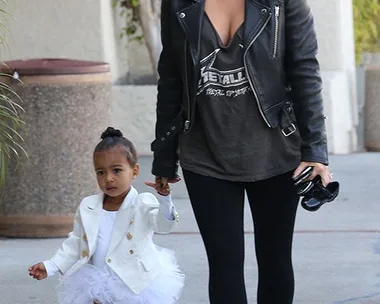 Woman in black leather jacket with toddler in white tutu and jacket, holding hands while walking outside.