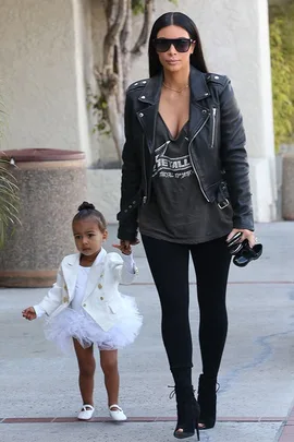 Woman in black leather jacket with toddler in white tutu and jacket, holding hands while walking outside.