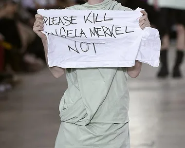 A model on runway holds a sign reading "PLEASE KILL ANGELA MERKEL NOT" at Rick Owens fashion show.