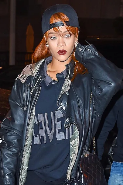 Rihanna wearing a cap, leather jacket, and "CAVE" sweatshirt, with bold lipstick, posing outdoors at night.