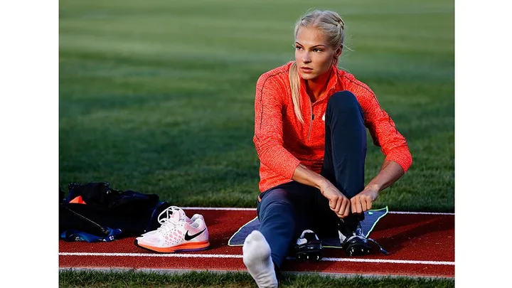 Woman in a red jacket sitting on a track, adjusting her shoe, with sports gear next to her on the grass.