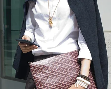 A person in a white shirt and dark coat, holding a patterned bag, looking at their phone.