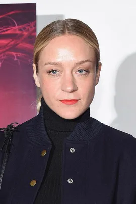 Chloë Sevigny in a dark jacket and turtleneck, standing against a background with red and purple lighting effects.