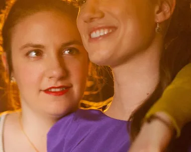 Two women, one in a purple dress smiling with her arm around the other, in warm lighting.