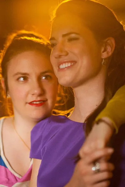 Two women, one in a purple dress smiling with her arm around the other, in warm lighting.