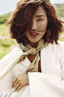 "Person with short brown hair wearing a white blazer and scarf, in a field with flowers and greenery in the background."