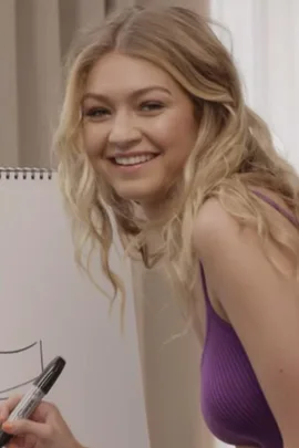 Woman drawing on paper while smiling, wearing a purple tank top; playing Pictionary.