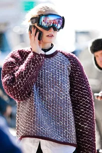 A woman in a textured sweater and ski goggles talks on a mobile phone outdoors in winter.