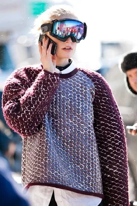 A woman in a textured sweater and ski goggles talks on a mobile phone outdoors in winter.