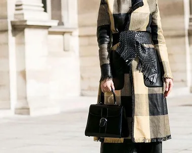 Woman in a plaid coat with a belt, wearing sunglasses, holding a black handbag, standing outside in a sunny setting.