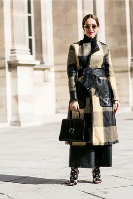 Woman in a plaid coat with a belt, wearing sunglasses, holding a black handbag, standing outside in a sunny setting.