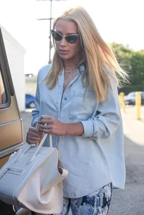 I don't know who this is. Woman with long blonde hair, wearing sunglasses, denim shirt, holding a beige handbag, standing outside with soft sunlight.
