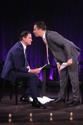 Two men in suits act out a skit on stage, holding papers, with one standing and leaning forward while the other is seated.