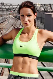 Woman in a neon yellow sports bra and gray workout pants standing on a bridge, with train tracks in the background.