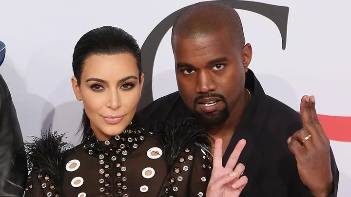 Kim Kardashian and Kanye West posing together, both showing hand gestures and dressed in black outfits with detailed designs.