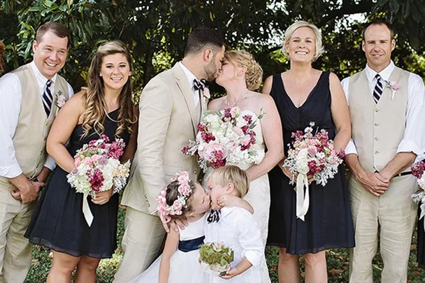 Wedding party photo with a couple kissing, the bridal party smiling, and a flower girl and ring bearer also sharing a kiss.