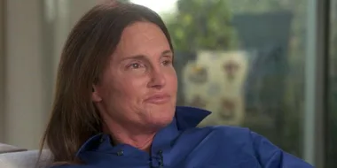 Caitlyn Jenner in a blue shirt, looking thoughtfully to the side, in a well-lit room with a blurred background.