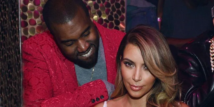 Kim Kardashian sitting while Kanye West, in a red jacket, looks at her smiling.