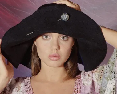 Young woman in a large black hat and lace outfit holding the hat's brim with both hands, posed against a textured background.