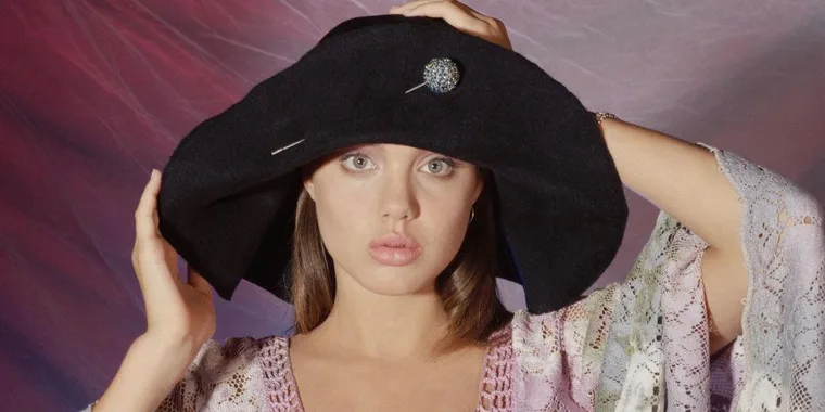 Young woman in a large black hat and lace outfit holding the hat's brim with both hands, posed against a textured background.