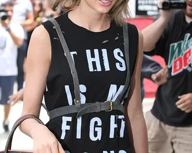 A woman wearing sunglasses, a black shirt with "THIS IS MY FIGHT SONG" text, a harness, and carrying a Louis Vuitton bag.