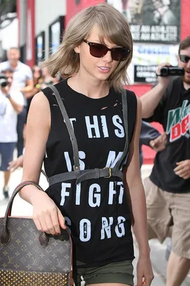 A woman wearing sunglasses, a black shirt with "THIS IS MY FIGHT SONG" text, a harness, and carrying a Louis Vuitton bag.