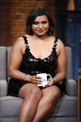 Mindy Kaling sitting on a talk show set, wearing a black dress and holding a mug.