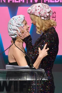 Nicole Kidman and Naomi Watts embrace on stage, both wearing decorative shower caps, at a media event.