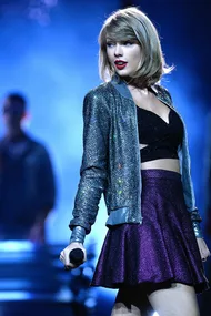 Taylor Swift performing on stage, wearing a glittery jacket and purple skirt.