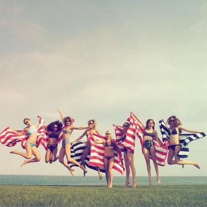 A group of women in swimsuits jump with striped towels, celebrating on a grassy area by the sea.