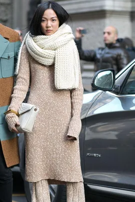 Woman in beige knit dress and scarf holding a quilted purse, standing next to a car on a city street.