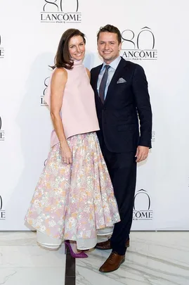 A man and a woman pose at the Lancôme 80th anniversary event; she wears a floral skirt, he wears a suit.