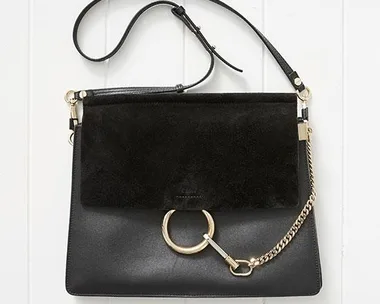 A black leather handbag with a suede flap, gold ring and chain detail, and an adjustable strap, on a white background.