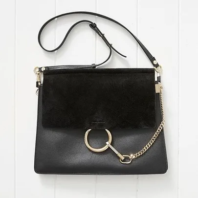 A black leather handbag with a suede flap, gold ring and chain detail, and an adjustable strap, on a white background.