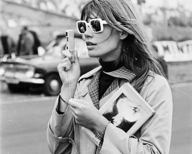 Woman with sunglasses and a trench coat holds a camera and a book at an outdoor event. Black and white photo.