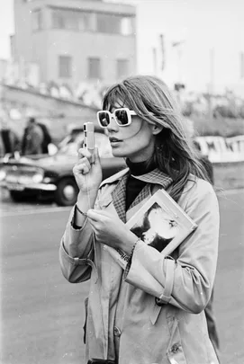 Woman with sunglasses and a trench coat holds a camera and a book at an outdoor event. Black and white photo.