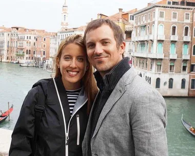 A smiling couple poses in front of a canal and historic buildings, with a gondola in the background.