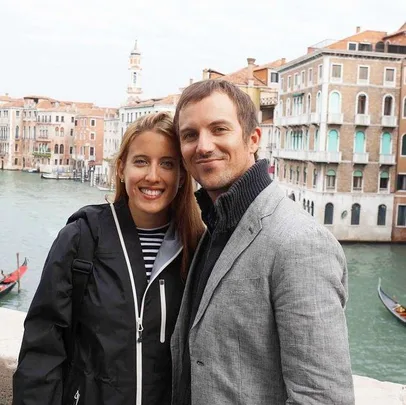 A smiling couple poses in front of a canal and historic buildings, with a gondola in the background.