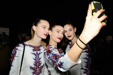 Three women with vibrant makeup, wearing matching patterned tops, take a selfie together in dark surroundings.